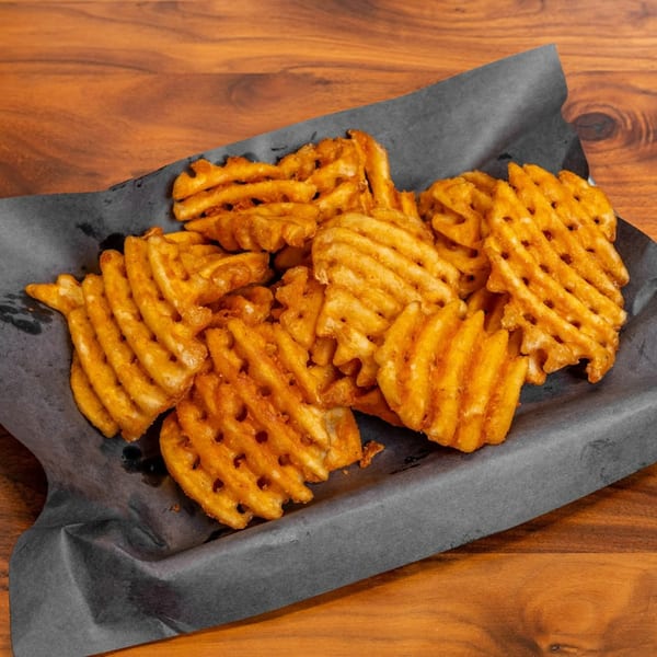 A close-up of golden-brown waffle fries served on dark paper, against a wooden table.